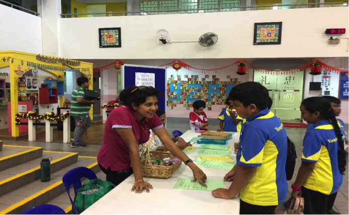 A woman in a red shirt leans over a table helping children in blue and yellow uniforms at a school event.